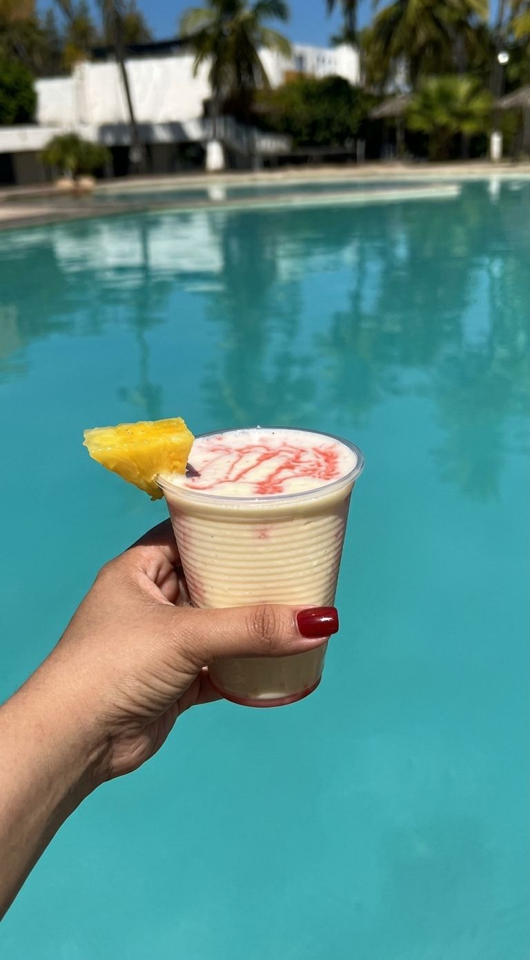 Eine Hand, die ein Getränk mit Ananas-Garnitur hält, vor einem Pool mit klarem, blauem Wasser.