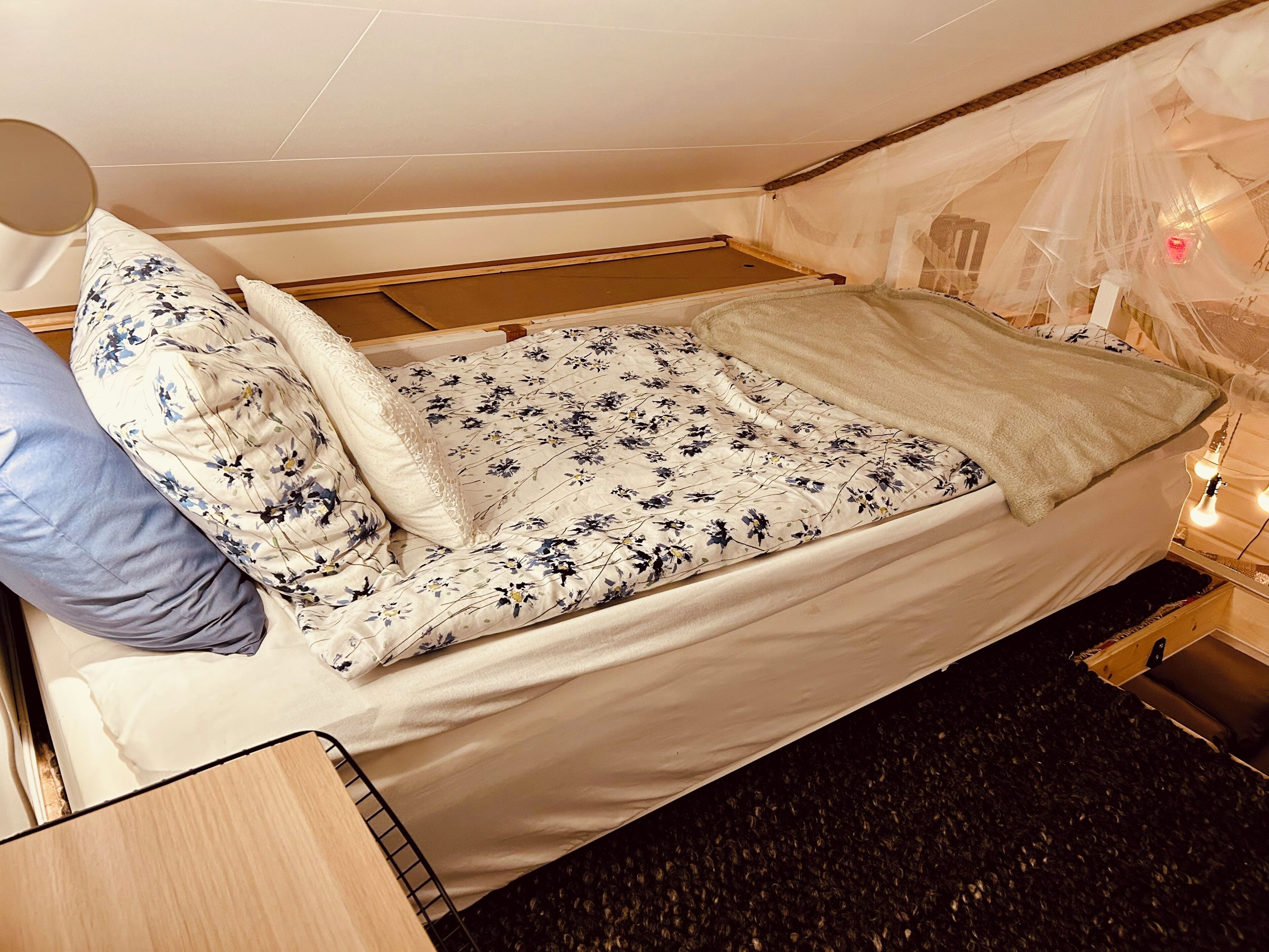 A single bed with a floral bedspread, a white pillow, and a blue pillow. There is a bedside table with a lamp and a small shelf.