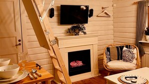 Ett rum i loftstil med en trätrappa, en tv som är monterad på väggen, en öppen spis, ett matbord med bestick och en fåtölj i rotting.