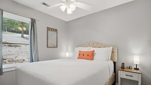 Quarto com cama, mesa de cabeceira, ventoinha no teto, janela com cortinas e espelho na parede.