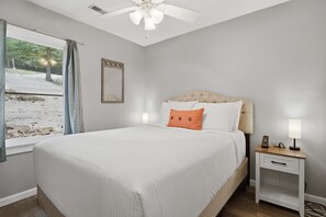 Ein Schlafzimmer mit einem Bett, einem Nachttisch, einem Deckenventilator, einem Fenster mit Vorhängen und einem Spiegel an der Wand.