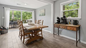 Uma área de refeições com mesa e cadeiras de madeira, um espaço para café e vista para o verde através de grandes janelas.