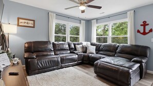Uma sala de estar com sofá modular, ventilador de teto e grandes janelas.
