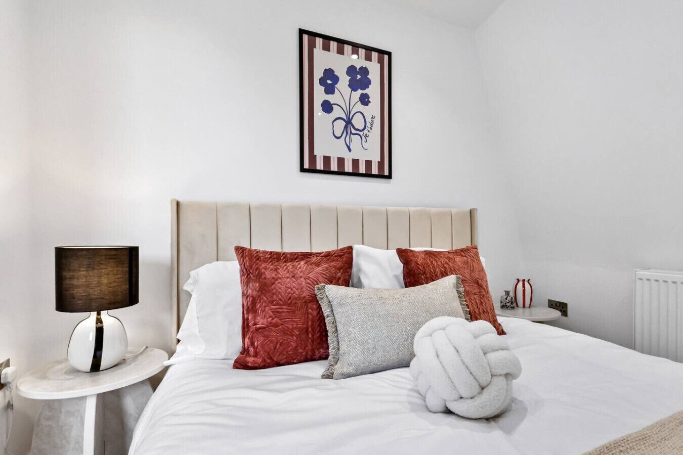 A neatly made bed with white bedding, red and grey pillows, and a decorative pillow with a bow. A bedside lamp and a framed artwork are visible on the wall.