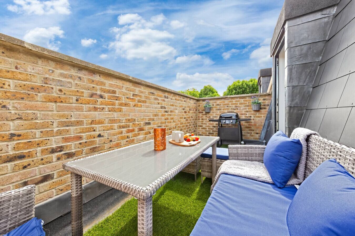 An outdoor patio with a brick wall, a table with a plate of food, a grill, and a wicker sofa with cushions.