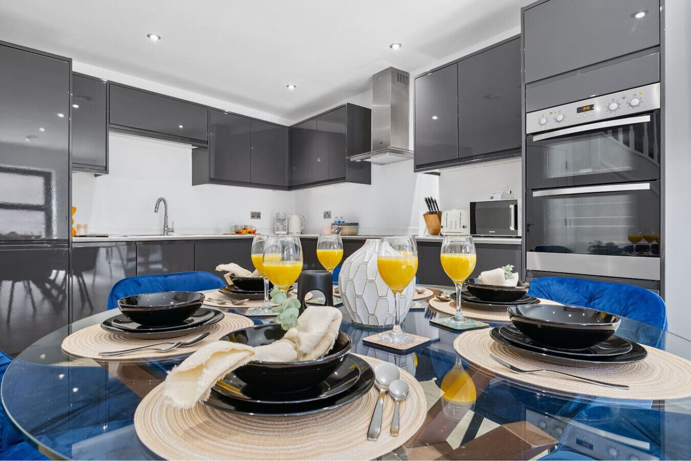 A modern kitchen with a dining area, featuring a glass table, dark cabinets, and a built-in oven.