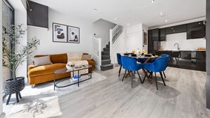 Uma sala de estar moderna com sofá, mesa de centro e cadeiras azuis. Há uma escadaria que leva a uma cozinha com área de jantar.