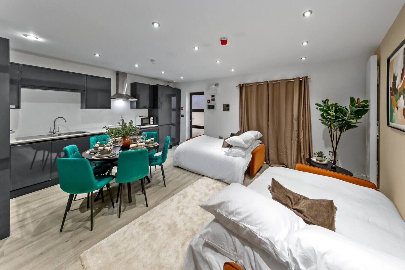 Una habitación de hotel moderna con kitchenette, zona de comedor, dos camas y un ventanal con cortinas.