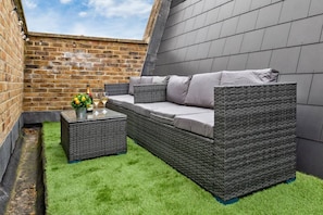 A corner patio with a grey wicker sofa, a small table with a bottle and glasses, and a brick wall. - Luxurious 5 BED | Sleeps 14 | Tower Bridge (Greater London)