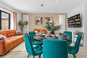 Ein modernes Wohnzimmer mit einer Couch, smaragdgrünen Sesseln, einem Esstisch für vier Personen und Kunstwerken an den Wänden.
