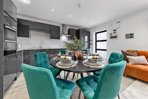 Une cuisine moderne avec des meubles de couleur sombre, une table à manger pour quatre personnes entourée de chaises turquoise et un canapé dans l’espace salon.