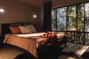 A bedroom with a large window, a bed with a brown blanket, and a rug on the floor.