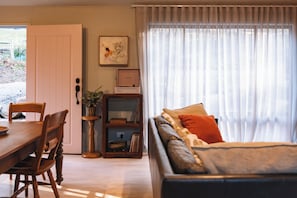 A living room with a wooden dining table, a sofa with cushions, a bookshelf, and a window with sheer curtains.