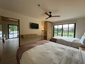 Ein Hotelzimmer mit zwei Betten, einem Deckenventilator, einem an der Wand montierten Fernseher und großen Fenstern mit Blick ins Freie.