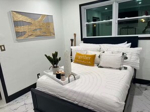 Ein ordentlich eingerichtetes Schlafzimmer mit einem Bett, einem Nachttisch, einem Fenster und einem an der Wand hängenden abstrakten Kunstwerk.