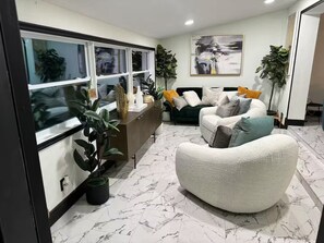 Ein modernes Wohnzimmer mit einer Couch, einem Couchtisch und einer Anrichte. Es gibt Blumentöpfe und ein Bild an der Wand.