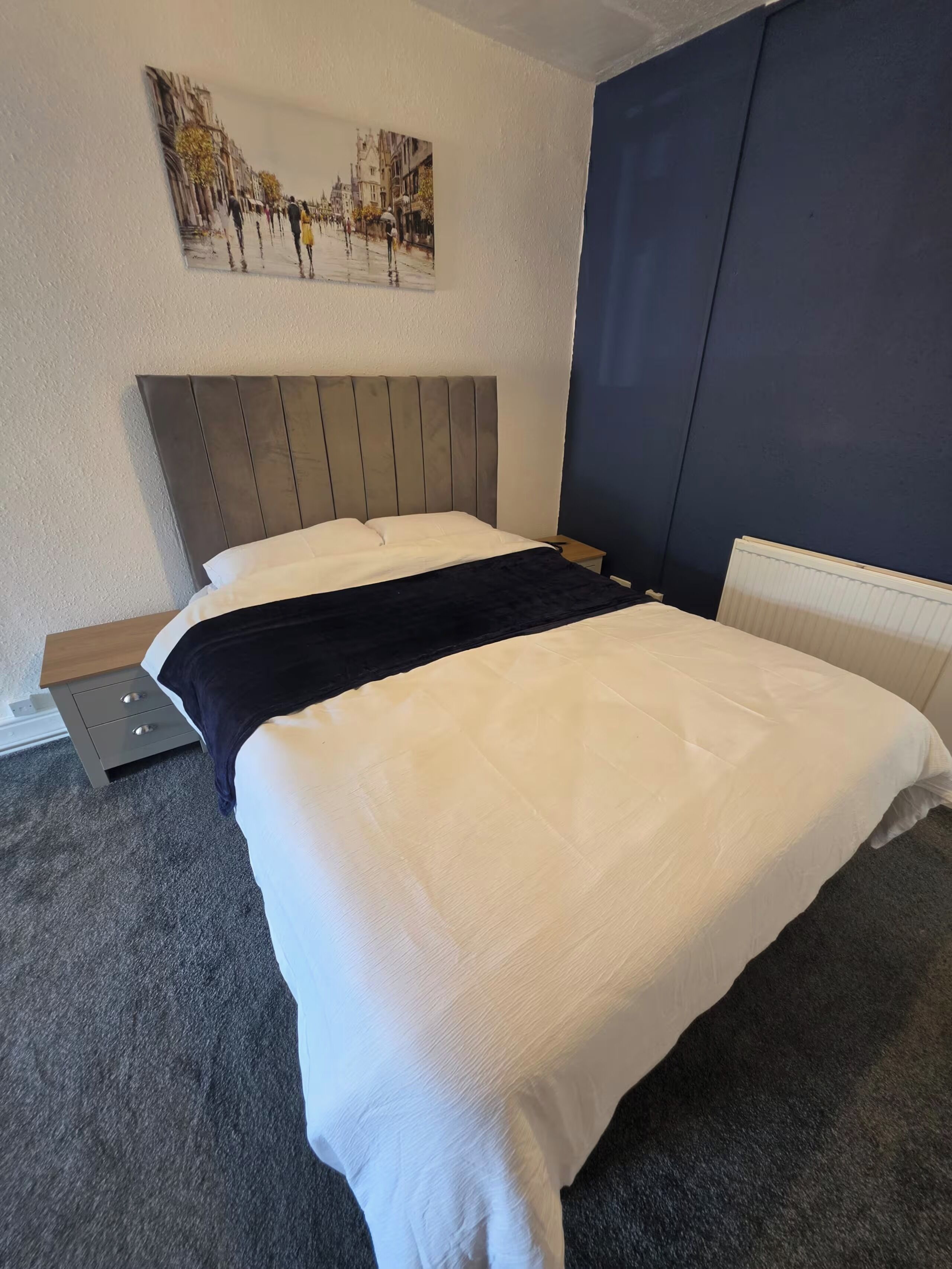 Ein Hotelzimmer mit einem Bett, einem Nachttisch, einer Lampe, einem Bild an der Wand und einer dunkel gefärbten Wand.