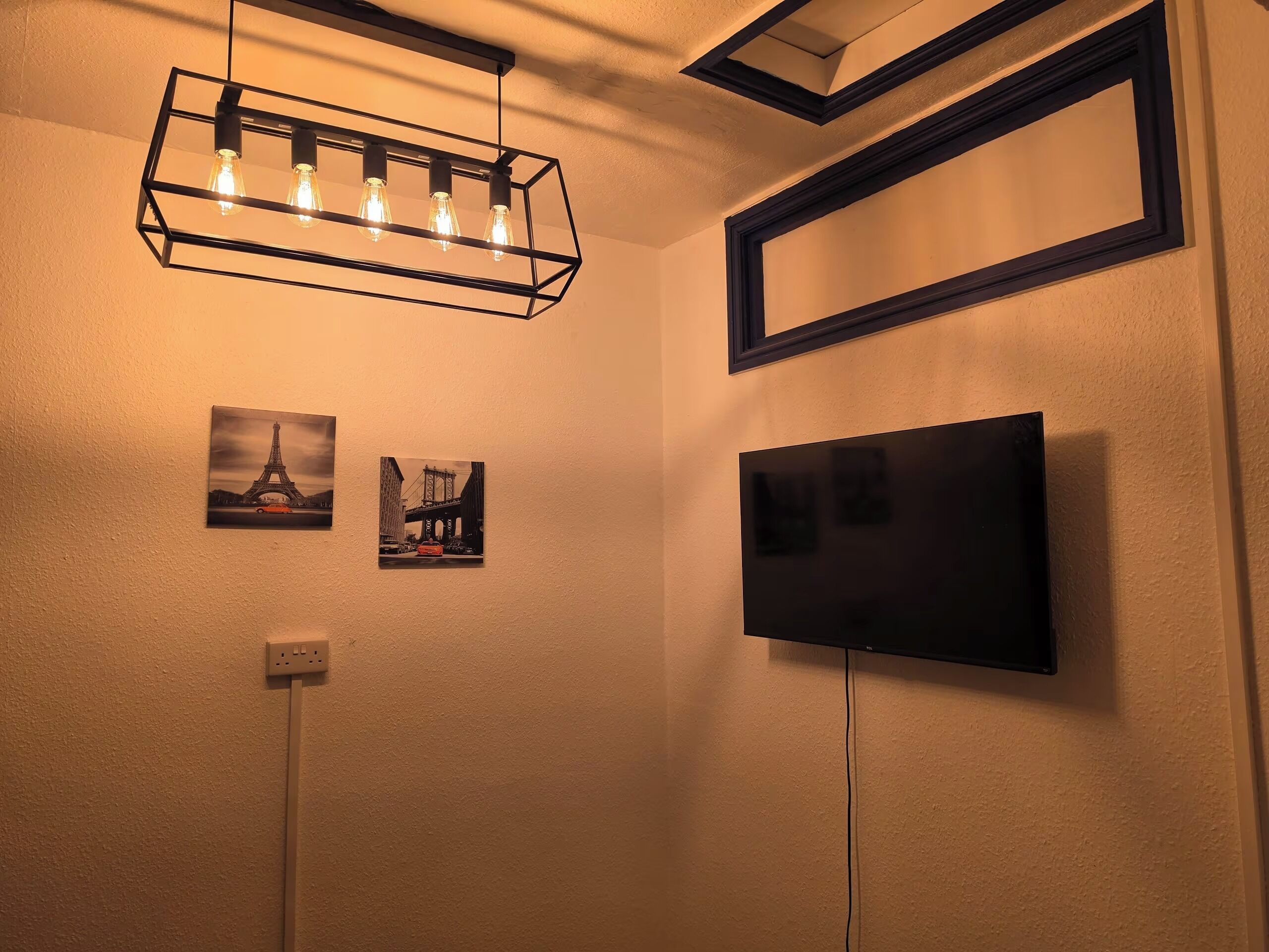 Ein Zimmer mit flachbildfernseher an der Wand, einer schwarzen Leuchte mit freiliegenden Glühbirnen und zwei gerahmten Bildern von Brücken.