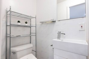 A bathroom with a white sink, toilet, and shower. There are shelves with plants and a mirror above the sink. - Peaceful 4-bedroom apartment with AC in vibrant Flushing (Flushing)