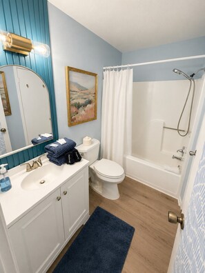 A bathroom with a white sink, toilet, and bathtub, a blue wall, and a painting. - Fully Renovated Beach Retreat Townhome Combo! (Panama City Beach)
