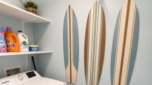 Una lavanderia con una lavatrice a carica frontale, scaffali con prodotti per la pulizia e tre decorazioni a forma di tavola da surf.