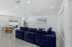A modern living room with a blue sectional sofa, a dining table with chairs, and a painting of a beach scene.