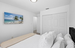 A bedroom with a bed, white walls, a door, and a painting of a beach scene.