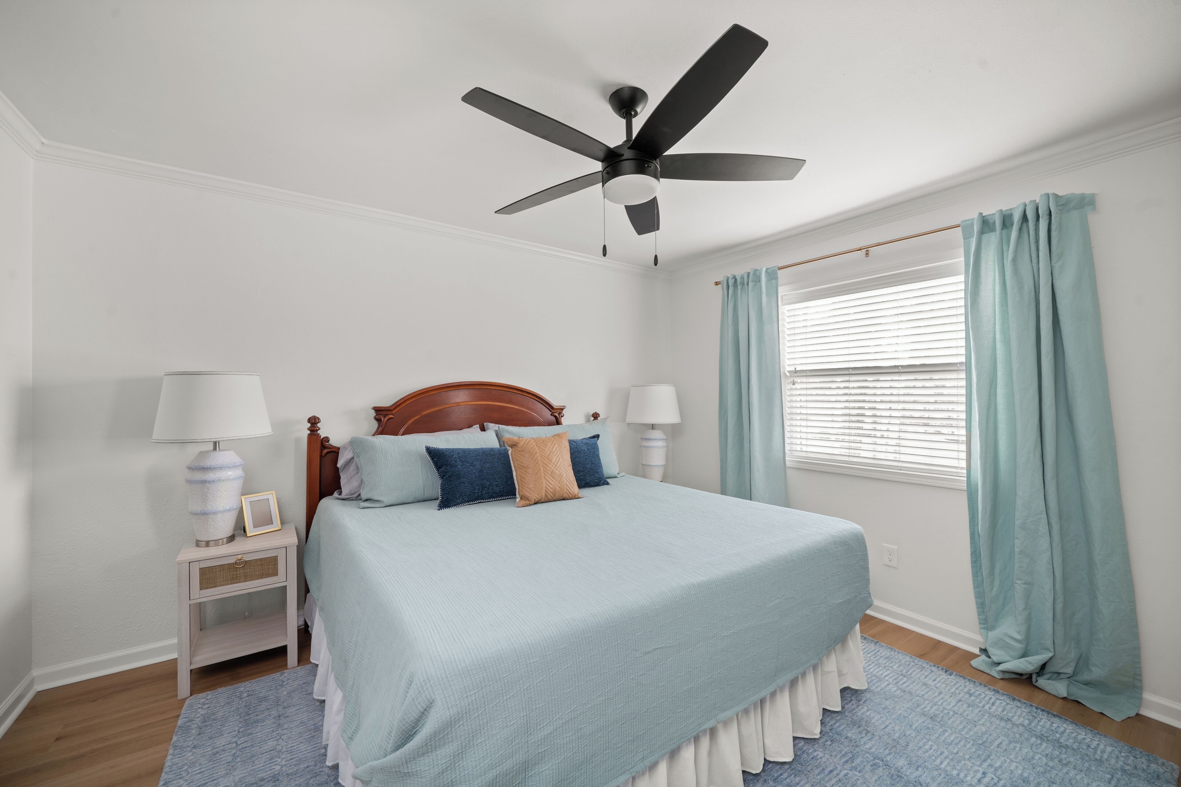 Une chambre à coucher avec un lit, un ventilateur de plafond, une table de chevet, une lampe, une fenêtre avec des rideaux et un plancher en bois.