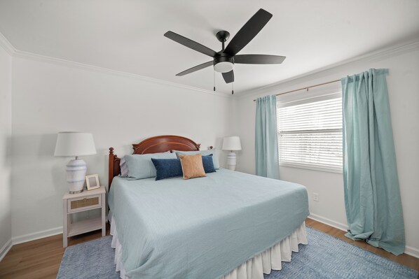 A bedroom with a bed, ceiling fan, nightstand, lamp, window with curtains, and a wooden floor.