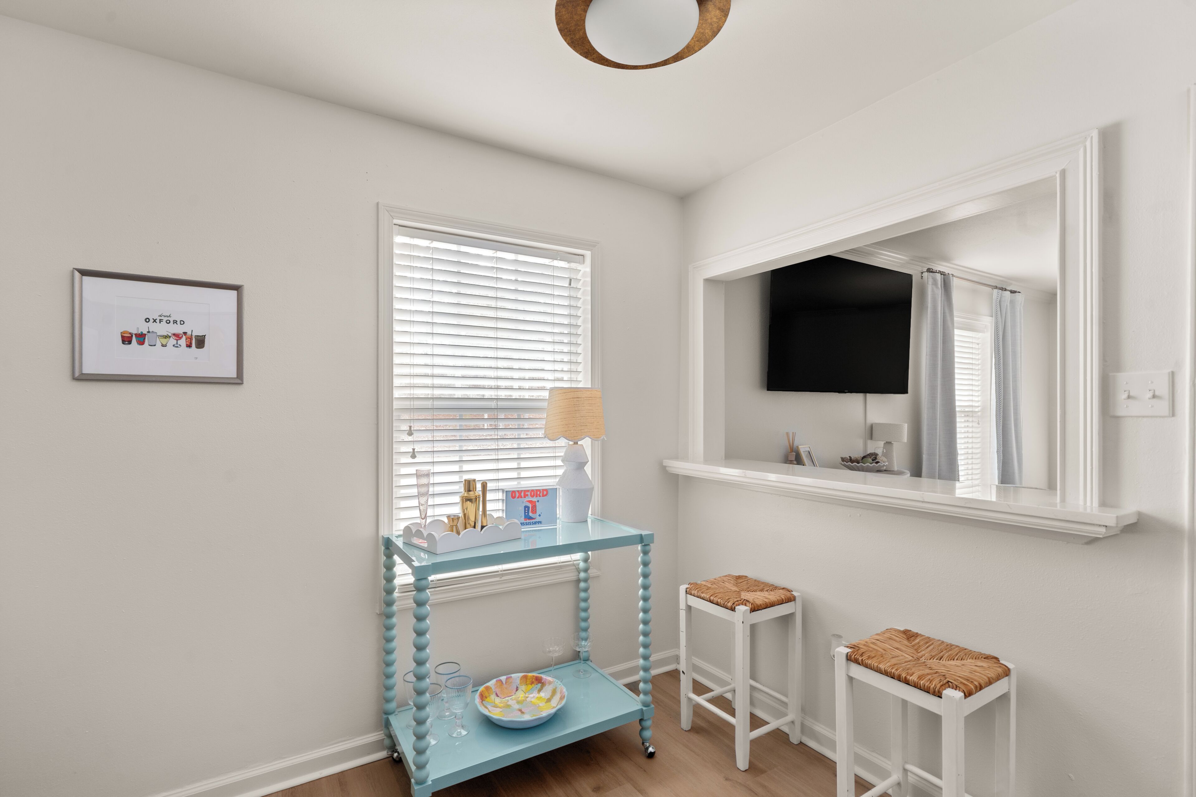 A room with a glass console table, two white stools, a framed picture on the wall, and a window with blinds.