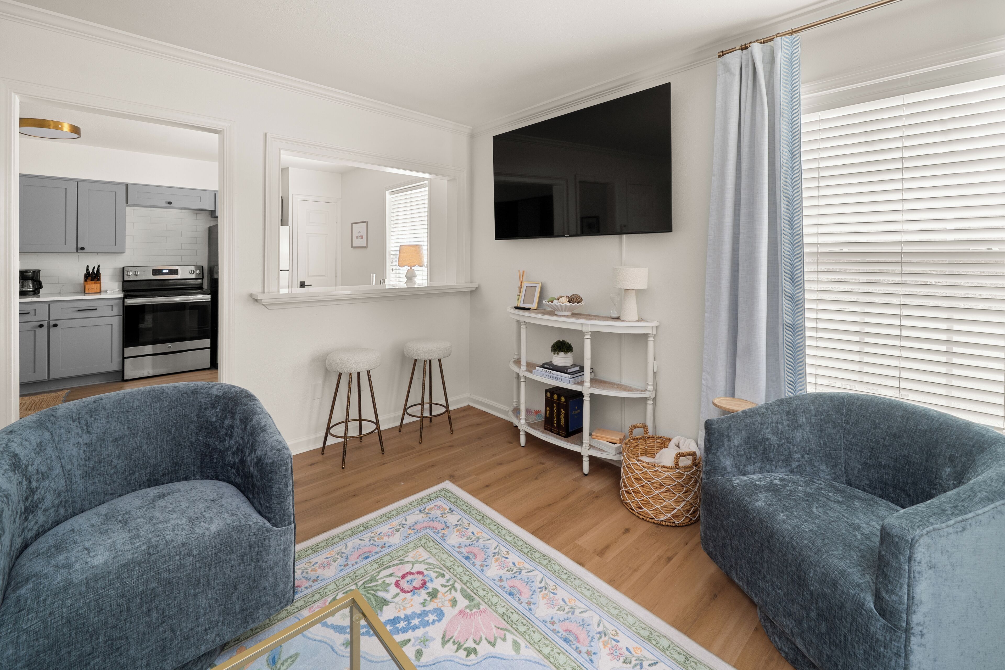 A modern living room with a flat-screen TV, two blue armchairs, a coffee table, a small white shelf with decorative items, and a kitchen area visible through an open door.