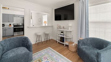 A modern living room with a flat-screen TV, two blue armchairs, a coffee table, a small white shelf with decorative items, and a kitchen area visible through an open door.
