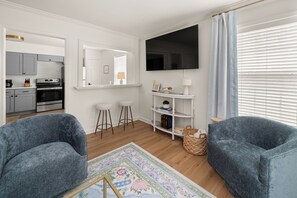 A modern living room with a flat-screen TV, two blue armchairs, a coffee table, a small white shelf with decorative items, and a kitchen area visible through an open door.