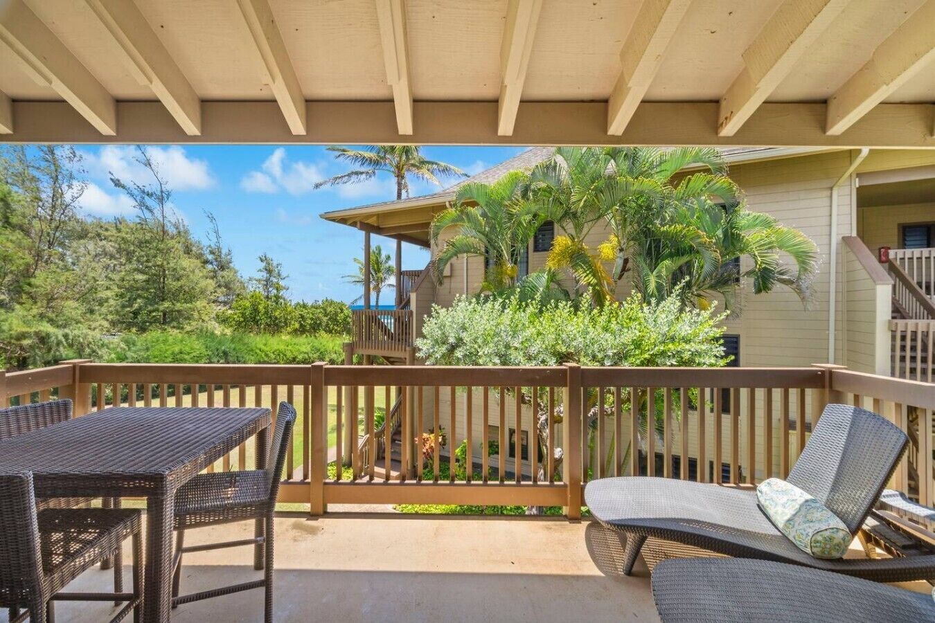 A covered outdoor area with a wooden deck, wicker furniture, and a view of a building with a palm tree.
