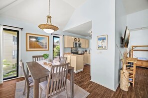 A modern kitchen with wooden cabinets, a dining area with a table and chairs, and a view through a glass door.