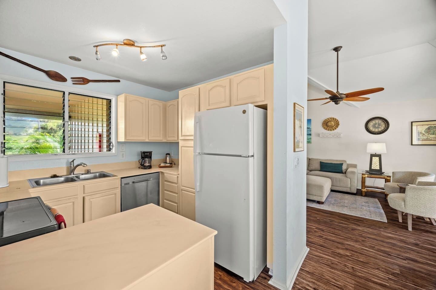 Cocina compacta con refrigerador, lavaplatos y lavamanos, zona de comedor con mesa, y sala de estar con sofá y ventilador de techo.