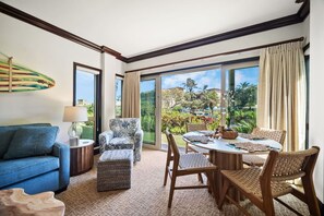 A room with a blue sofa, patterned armchair, and wicker chairs around a round table with a view of a garden and buildings. - Waipouli C102 / 2 King Beds / AC  / Pool (Kapaʻa)