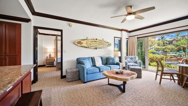 A living room with a blue sofa, a wooden coffee table, a ceiling fan, and a view of a tropical garden. - A/C Luxury Poolside Retreat / Resort fee included (Kapaʻa)