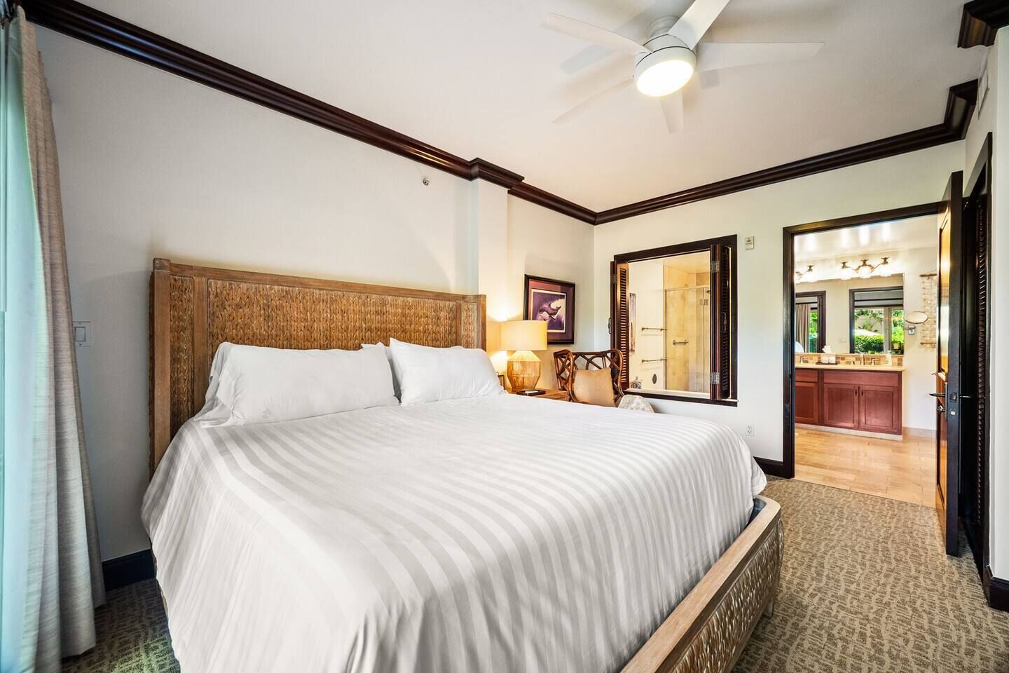 Habitación de hotel con una cama grande, ventilador de techo, mesita de noche con lámpara y vista al baño a través de una puerta abierta.