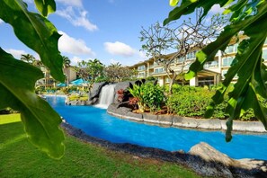 A resort pool with a waterfall feature, surrounded by lush greenery and a multi-story building with balconies. - A/C Luxury Poolside Retreat / Resort fee included (Kapaʻa)