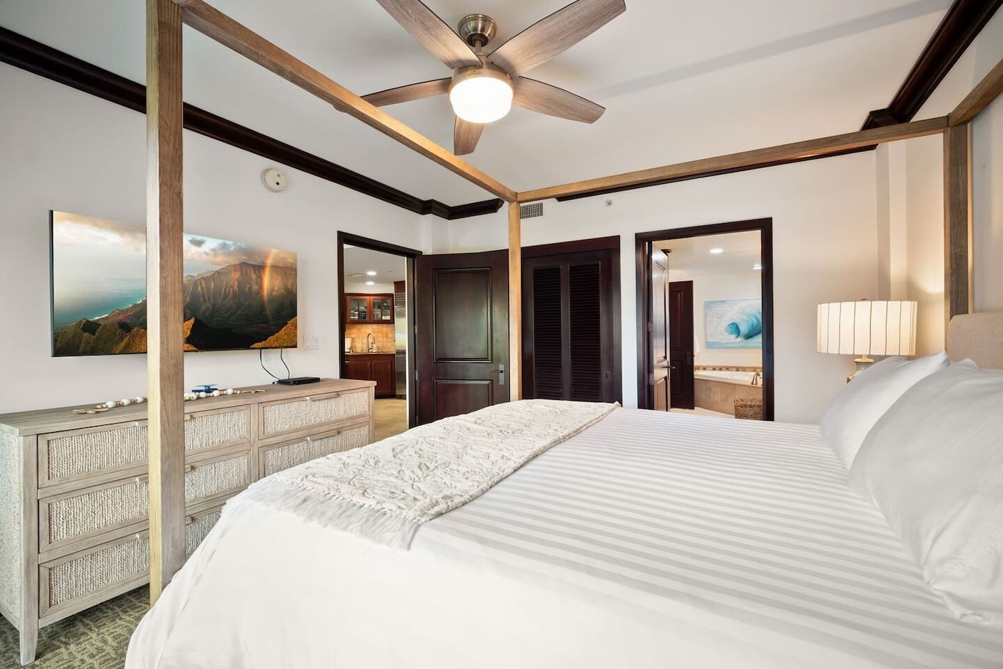 A hotel room with a large bed, a ceiling fan, a TV, and a view of the kitchen.