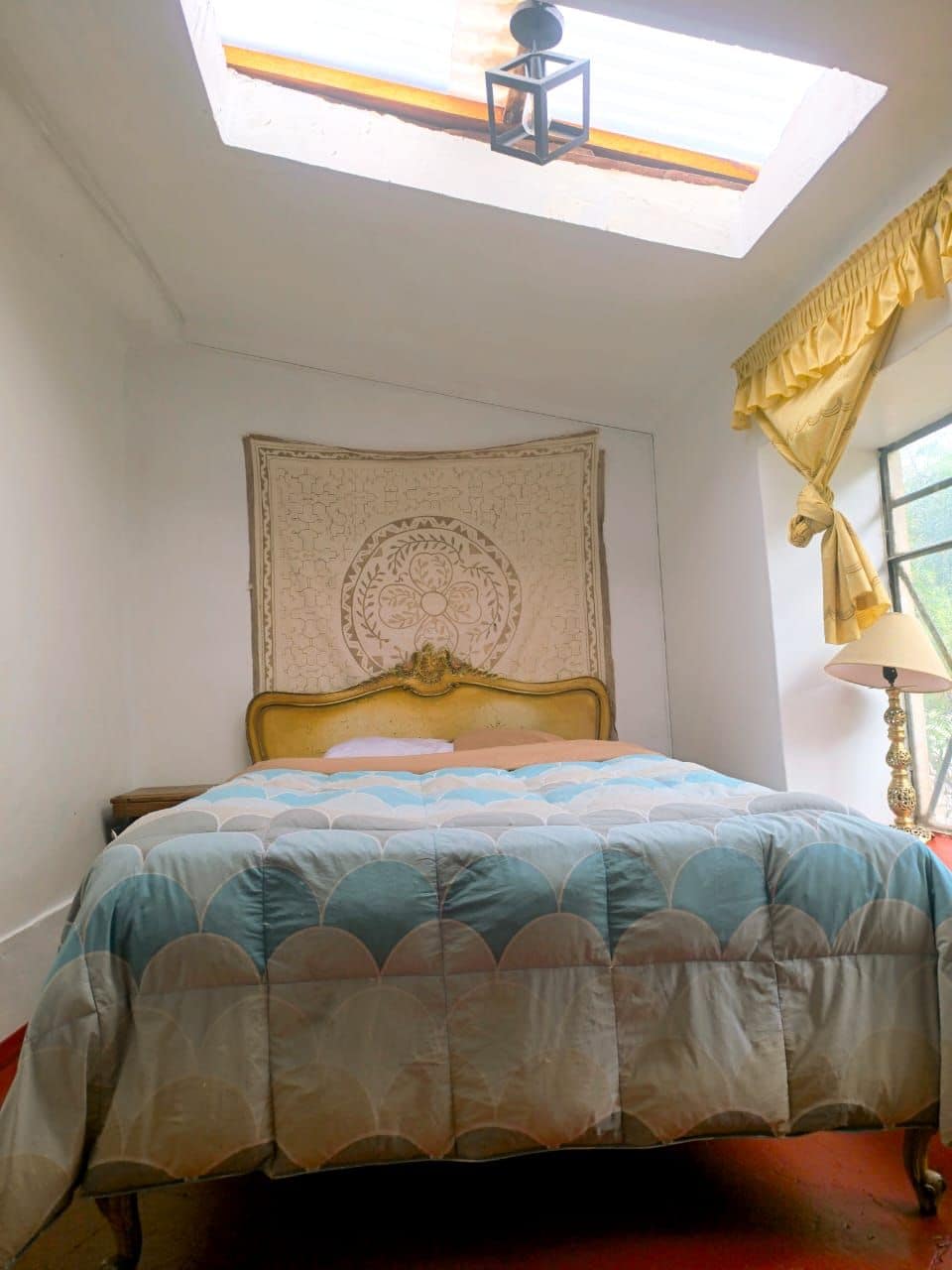 A bedroom with a bed, a headboard, a tapestry, a window with curtains, and a skylight.