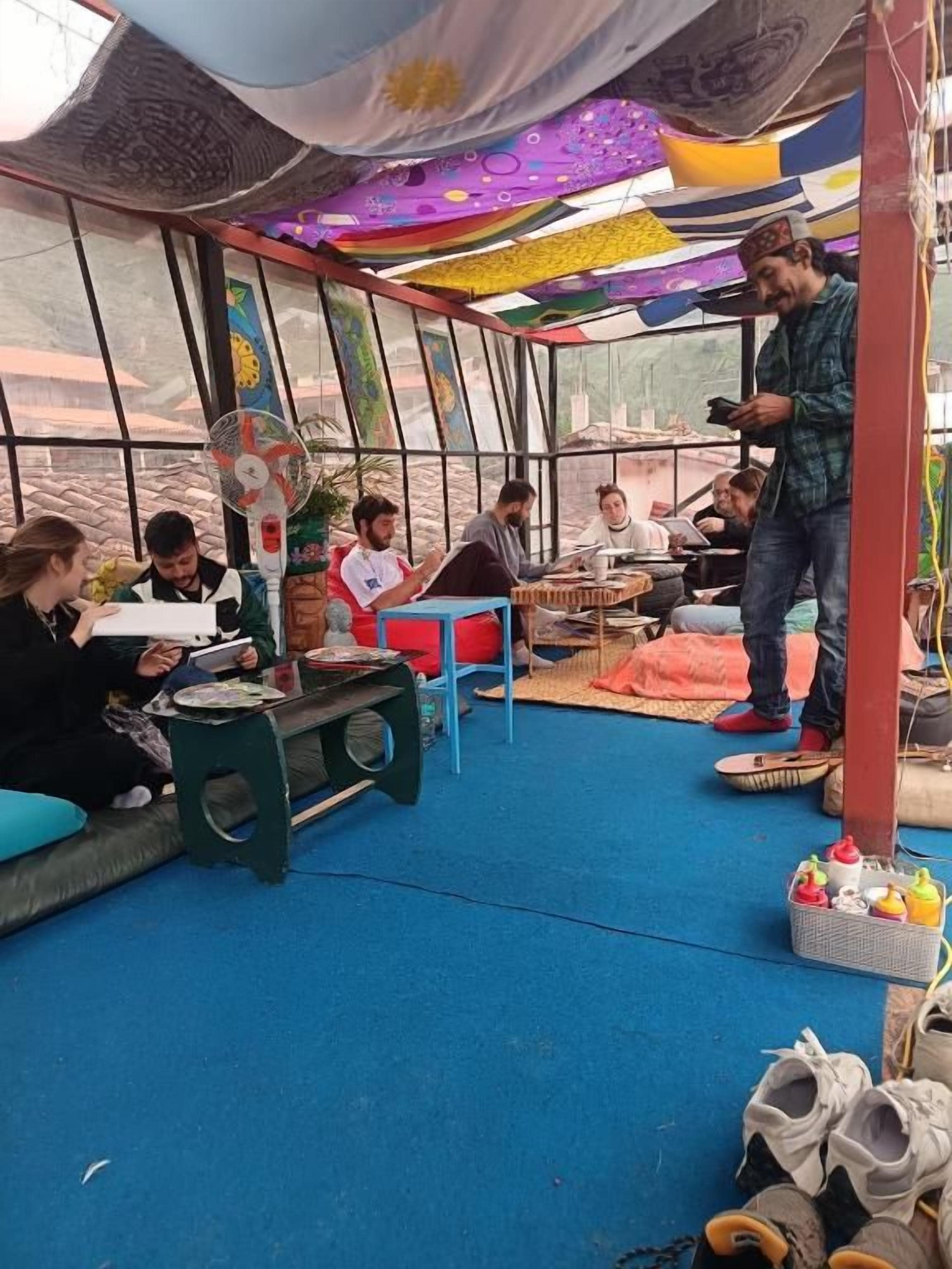 A group of people seated on benches inside a tent-like structure with a fan and a basket of items on the floor.