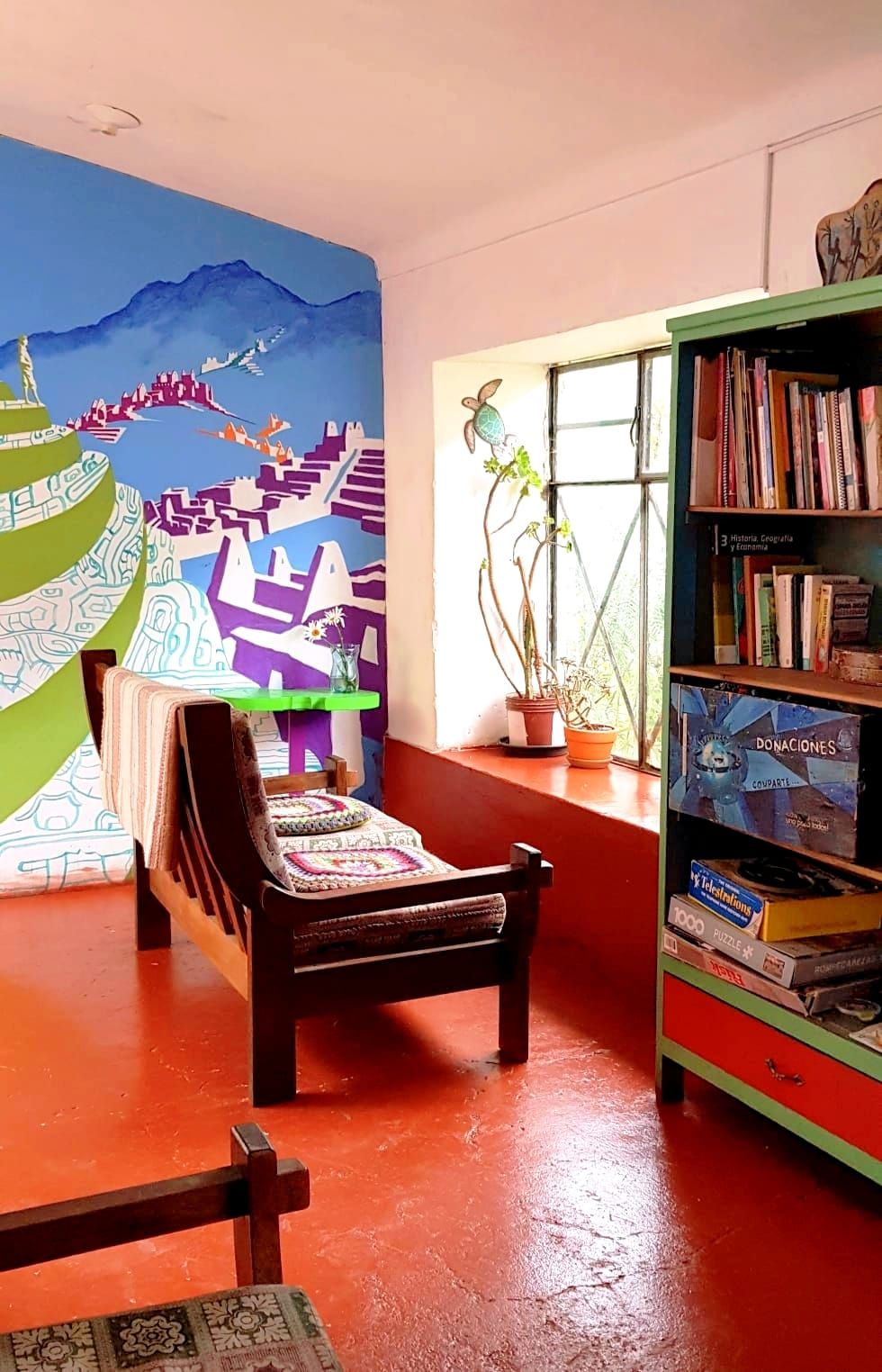 A room with a colorful mural, a wooden chair, a bookshelf, and a window with a plant.