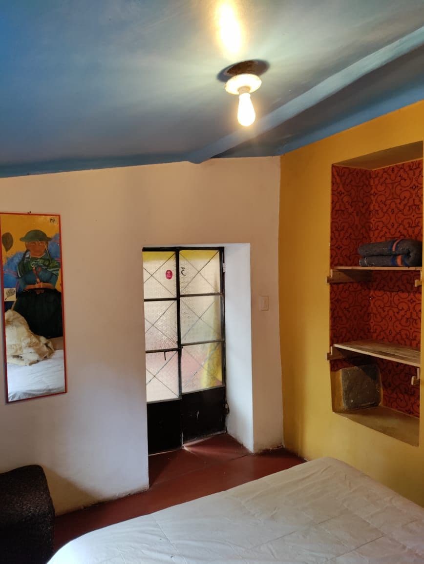 Une pièce avec un lit, une fenêtre à carreaux, une étagère avec des objets et un tableau au mur.