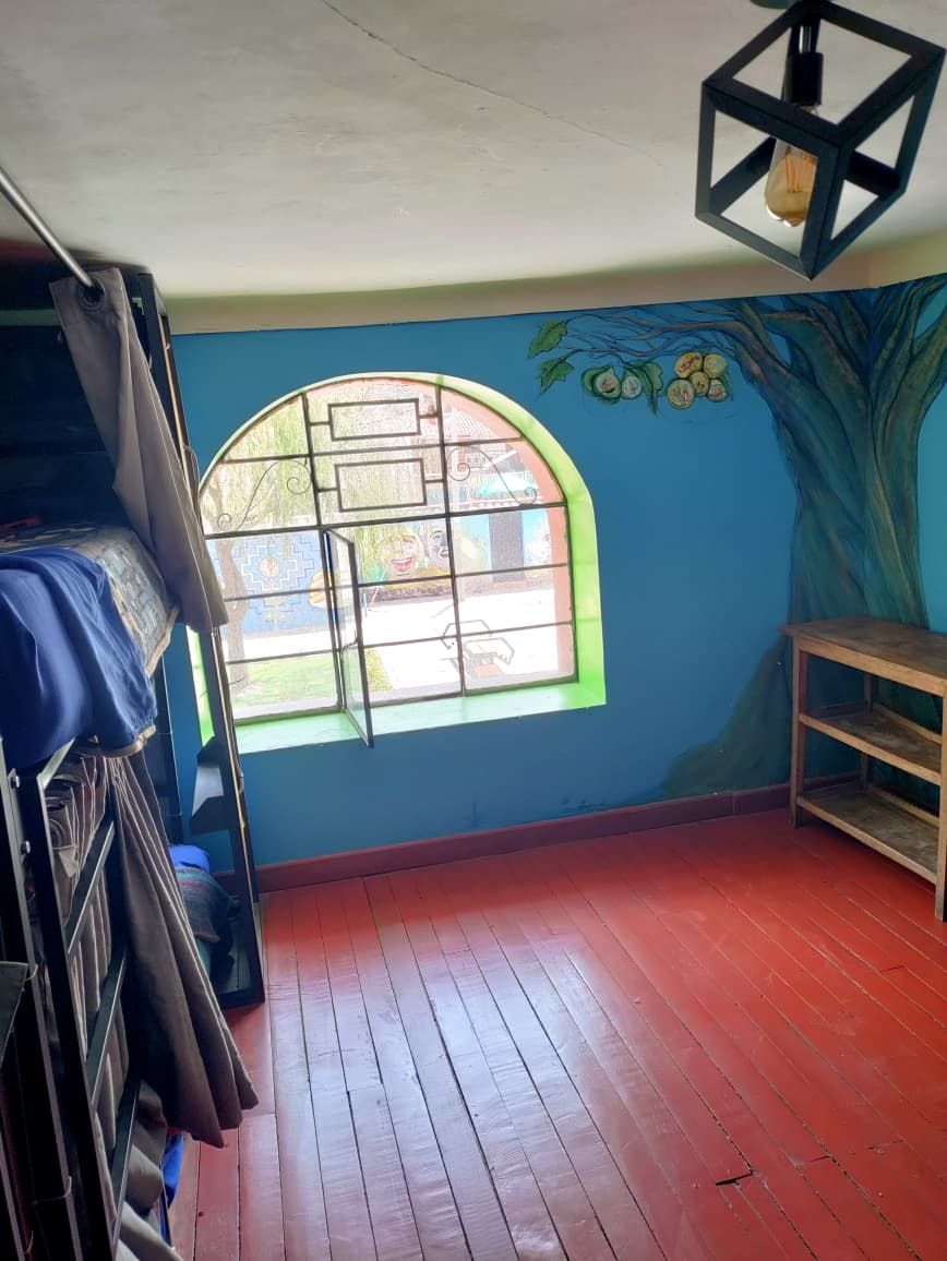 A room with a wooden floor, a window with a geometric design, a wall mural of a tree, and a hanging light fixture.