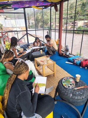 A group of people sitting on green and blue furniture in a room with large windows, a fan, and a Buddha statue. - Aguaymanto Hostel (Pisac)