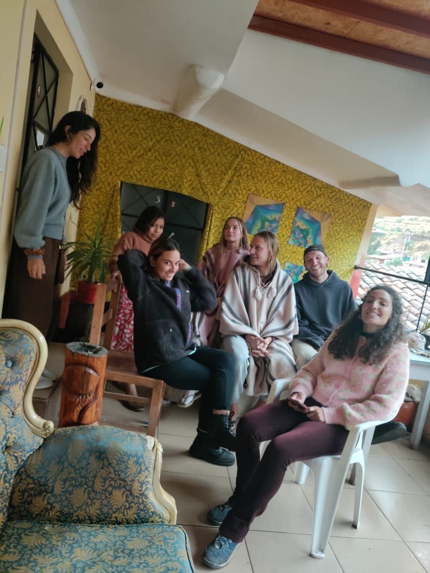 A group of people seated in a room with patterned wallpaper, a wooden chair, and a potted plant.