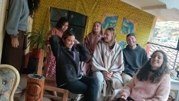 A group of people seated in a room with patterned wallpaper, a wooden chair, and a potted plant.