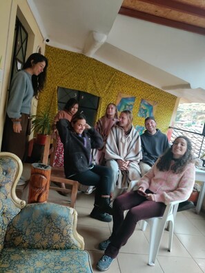 Un grupo de personas sentadas en una habitación con papel tapiz estampado, una silla de madera y una planta en maceta.